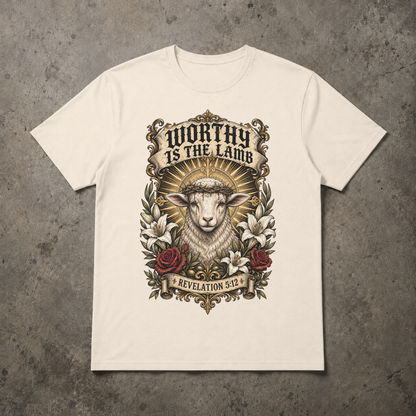 A ivory short-sleeve crewneck t-shirt with a detailed graphic print of a lamb wearing a crown of thorns, surrounded by lilies and roses, with the text 'WORTHY IS THE LAMB' and 'REVELATION 5:2'. The t-shirt is displayed flat on a textured gray concrete background.