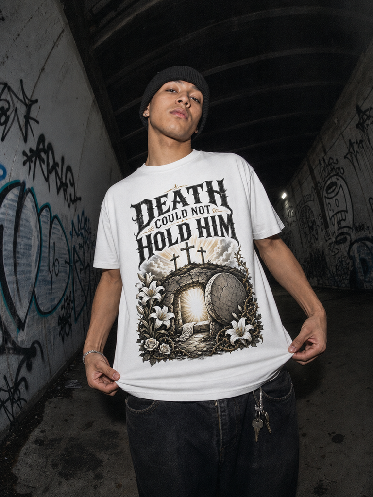 A man wearing a white graphic t-shirt with a gothic-style print that reads 'DEATH COULD NOT HOLD HIM' above an illustration of a tombstone, crosses, and white lilies. He is also wearing a black beanie and dark jeans, standing in a dark, graffiti-covered tunnel.
