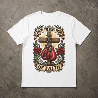 A white short-sleeve crewneck t-shirt with a detailed graphic print on the front, laid flat on a textured gray concrete background. The graphic features a wooden cross, red boxing gloves, red roses, and banners reading 'FIGHT THE GOOD FIGHT' and 'OF FAITH'.