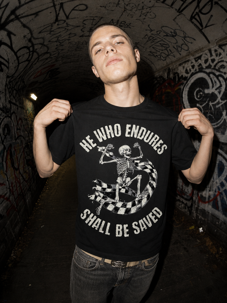 A man wearing a black graphic t-shirt with a white skeleton and the text 'HE WHO ENDURES SHALL BE SAVED', posing in a graffiti-covered tunnel.