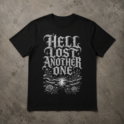 A black short-sleeve t-shirt with a large white gothic-style graphic reading 'HELL LOST ANOTHER ONE' above a floral and chain design, laid flat on a textured gray concrete background.