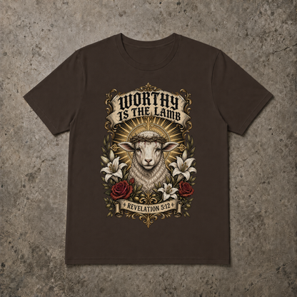 A brown short-sleeve crewneck t-shirt with a detailed graphic print of a lamb with crown of thorns, surrounded by lilies and roses, with the text 'WORTHY IS THE LAMB' and 'REVELATION 5:2'. The t-shirt is displayed flat on a textured gray concrete background.