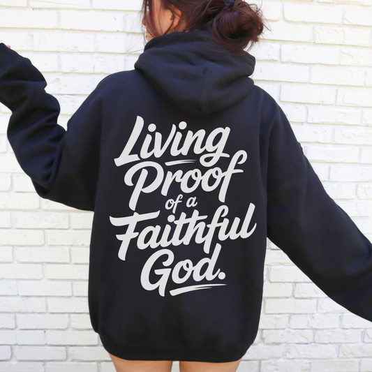 Person wearing a black christian streetwear hoodie with 'Living Proof of a Faithful God' text on the back against a white brick wall.