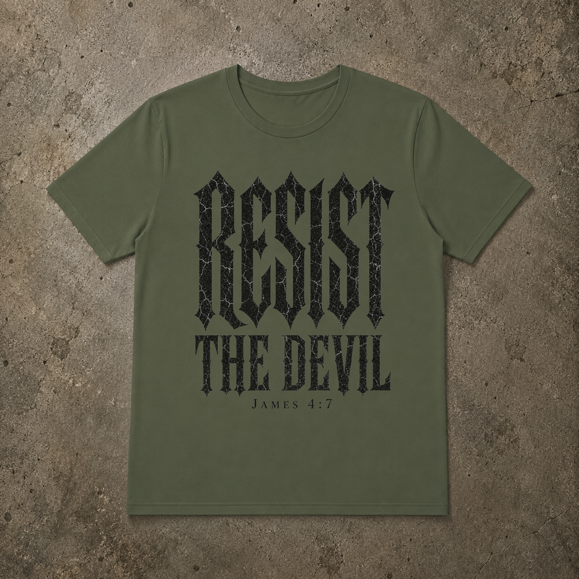 An olive green short-sleeve t-shirt with a distressed black graphic print reading 'RESIST THE DEVIL' and 'JAMES 4:7' underneath, laid flat against a textured concrete background.