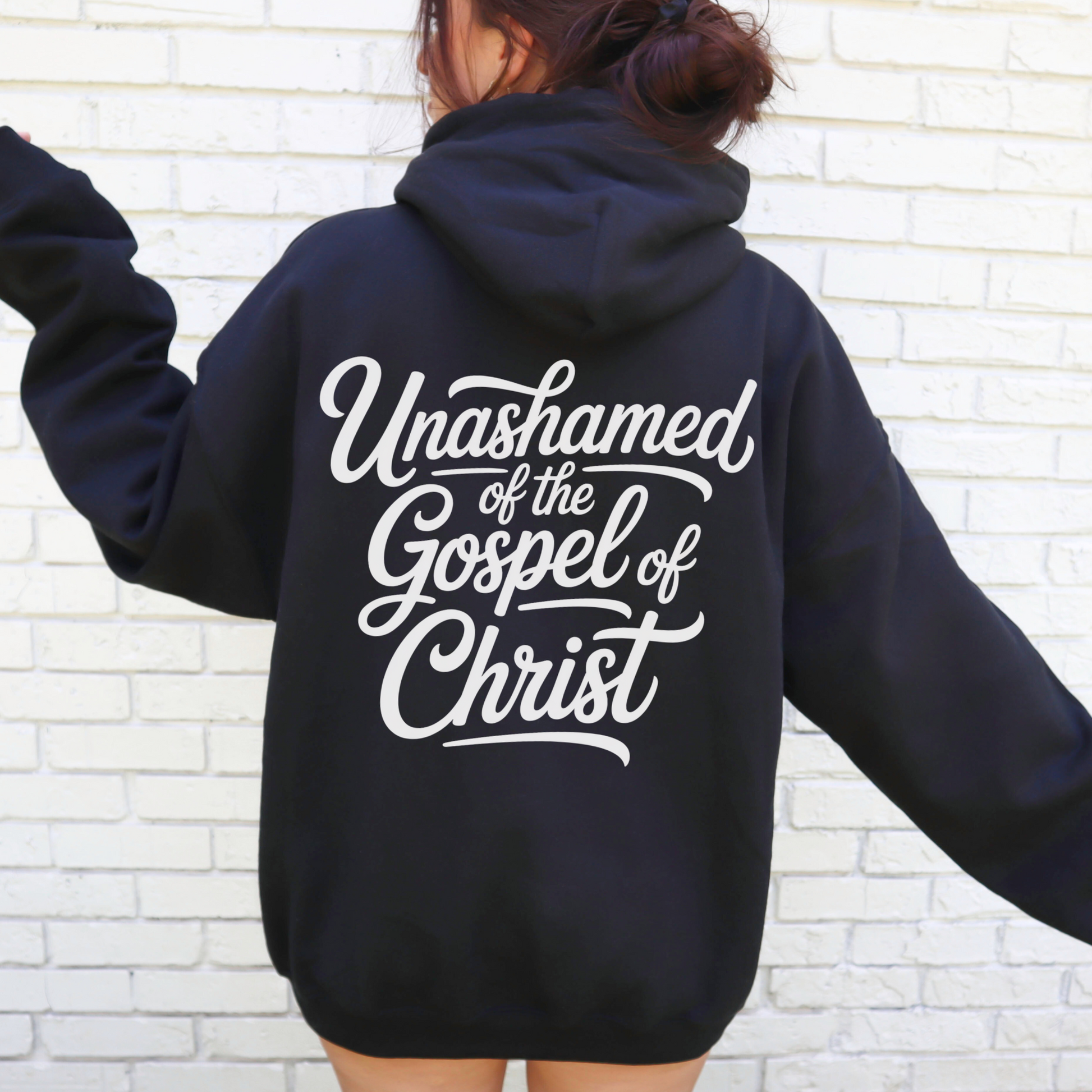 Person wearing a black gospel hoodie with 'Unashamed of the Gospel of Christ' text on the back against a white brick wall.